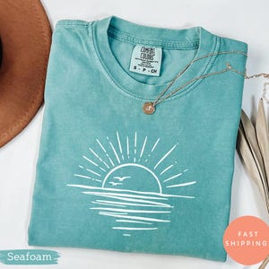 Op de afbeelding: Zeegroen Comfort Colors T-shirt met een witte afbeelding van een zon boven water en twee vogels. Het shirt heeft een ronde hals en korte mouwen. Een gouden ketting met een hanger ligt op het shirt. Het shirt is gemarkeerd met "S - P - CH".