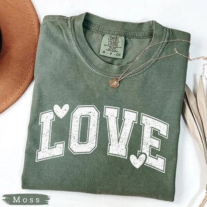 May include: A moss green t-shirt with the word "LOVE" in white block letters and two white hearts. The shirt has a crew neck and short sleeves. A gold necklace with a pendant is draped across the shirt. The word "Moss" is printed at the bottom.