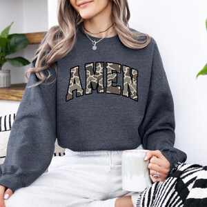May include: A dark gray sweatshirt with the word "AMEN" in a camouflage pattern across the chest. The sweatshirt has long sleeves and a crew neck. The person is holding a white mug.