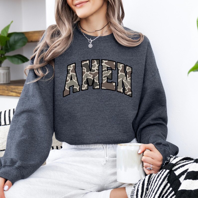 May include: A dark gray sweatshirt with the word "AMEN" in a camouflage pattern across the chest. The sweatshirt has long sleeves and a crew neck. The person is holding a white mug.