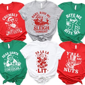 May include: Six Christmas-themed t-shirts in red, gray, green, and white. Each shirt features a unique graphic and text, including Santa Claus, gingerbread men, and festive phrases like "Crumby But Cute" and "Fa La La Lit."