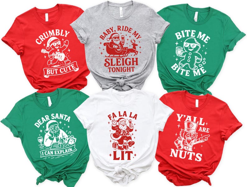 May include: Six Christmas-themed t-shirts in red, gray, green, and white. Each shirt features a unique graphic and text, including Santa Claus, gingerbread men, and festive phrases like "Crumby But Cute" and "Fa La La Lit."