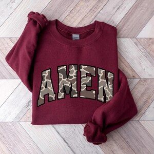 May include: A maroon sweatshirt with the word "AMEN" in a camouflage pattern across the chest. The sweatshirt is made of a soft material and has long sleeves. The garment is displayed on a wooden surface.