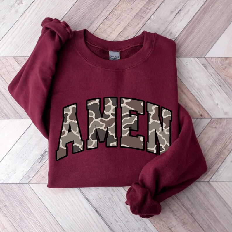 May include: A maroon sweatshirt with the word "AMEN" in a camouflage pattern across the chest. The sweatshirt is made of a soft material and has long sleeves. The garment is displayed on a wooden surface.