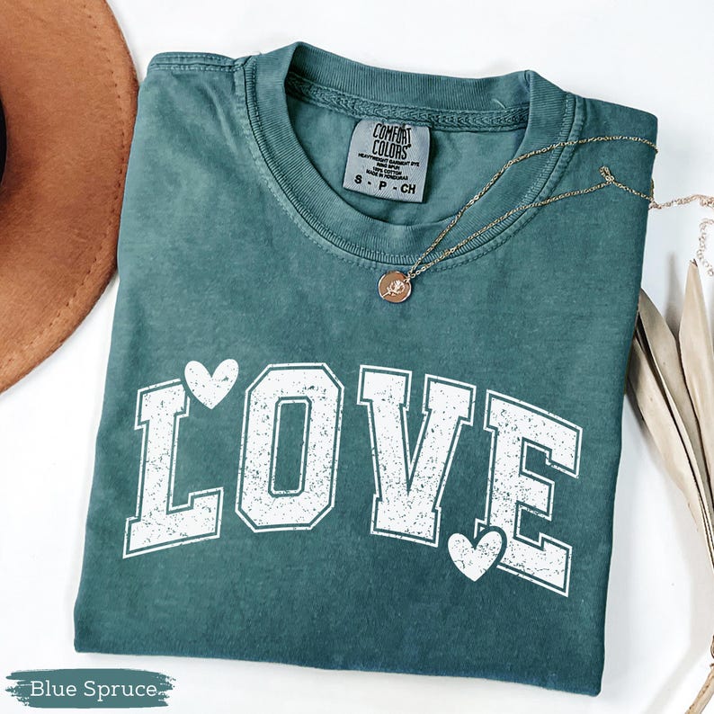 May include: Blue spruce t-shirt with the word "LOVE" in white block letters and two white hearts. A gold necklace with a pendant is on the shirt. The shirt is from Comfort Colors and is size S-P-CH.