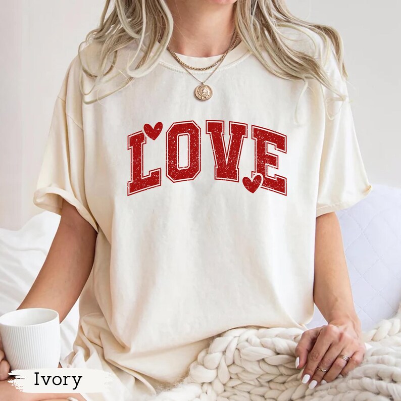 May include: Ivory-colored t-shirt with the word "LOVE" in red, collegiate-style lettering. Two small red hearts accent the design. The shirt has a relaxed fit and short sleeves. The word "Ivory" is printed at the bottom of the image.