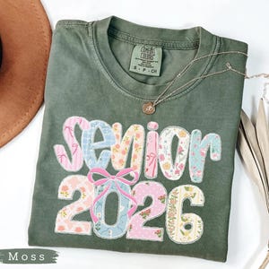 Może przedstawiać: Zielona koszulka Comfort Colors w kolorze mchu z napisem "Senior 2026" w patchworkowym wzorze. Patchwork zawiera kwiatowe i wzorzyste tkaniny w kolorze r&oacute;żowym, niebieskim i białym. Srebrny naszyjnik z wisiorkiem leży na koszulce.