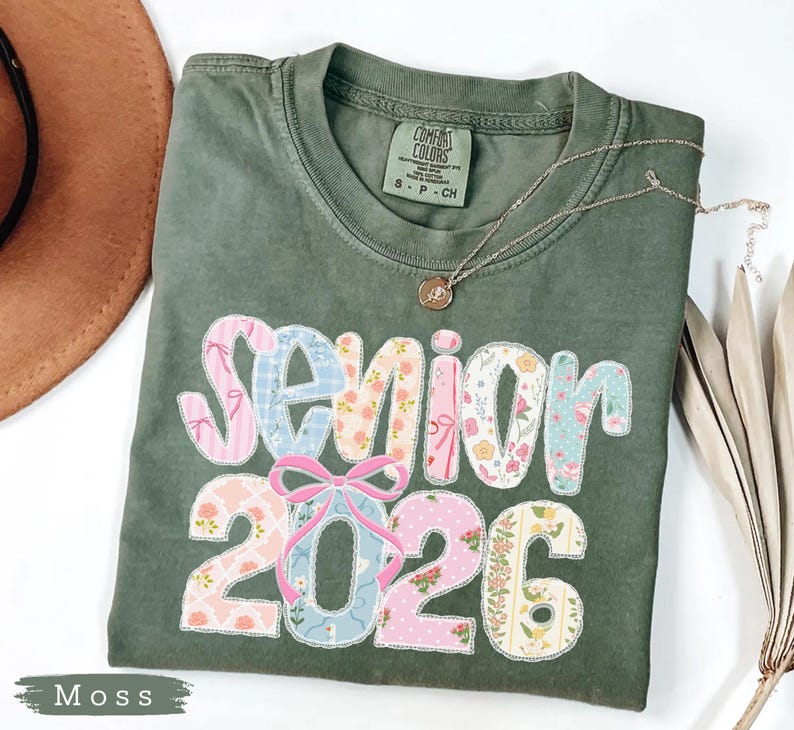 Może przedstawiać: Zielona koszulka Comfort Colors w kolorze mchu z napisem "Senior 2026" w patchworkowym wzorze. Patchwork zawiera kwiatowe i wzorzyste tkaniny w kolorze r&oacute;żowym, niebieskim i białym. Srebrny naszyjnik z wisiorkiem leży na koszulce.