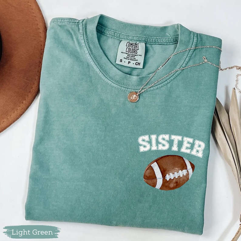 May include: Light green t-shirt with the word "SISTER" above a football graphic. The shirt has a crew neck and short sleeves. A gold necklace with a pendant is draped across the shirt.
