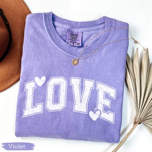 May include: A violet-colored t-shirt with the word "LOVE" printed in white, featuring heart accents. A gold necklace with a pendant rests on the shirt. The shirt is folded, and a brown hat and dried plant are in the background.