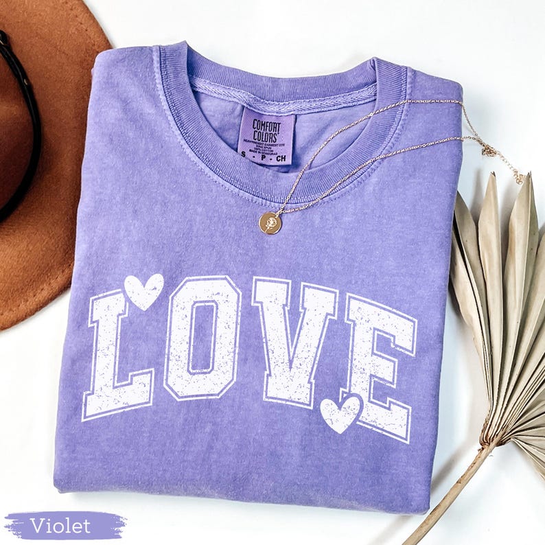 May include: A violet-colored t-shirt with the word "LOVE" printed in white, featuring heart accents. A gold necklace with a pendant rests on the shirt. The shirt is folded, and a brown hat and dried plant are in the background.