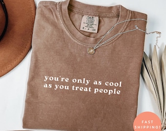 以下が含まれることがあります： 「you're only as cool as you treat people」の白文字がプリントされたエスプレッソカラーのTシャツ。シルバーのネックレスとペンダントがシャツの上にあります。シャツにはComfort Colorsのタグが付いています。