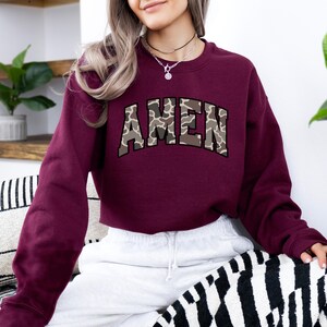 May include: A maroon sweatshirt with the word "AMEN" in a camouflage pattern across the chest. The sweatshirt has long sleeves and a crew neck. The person is wearing a silver necklace and white sweatpants.