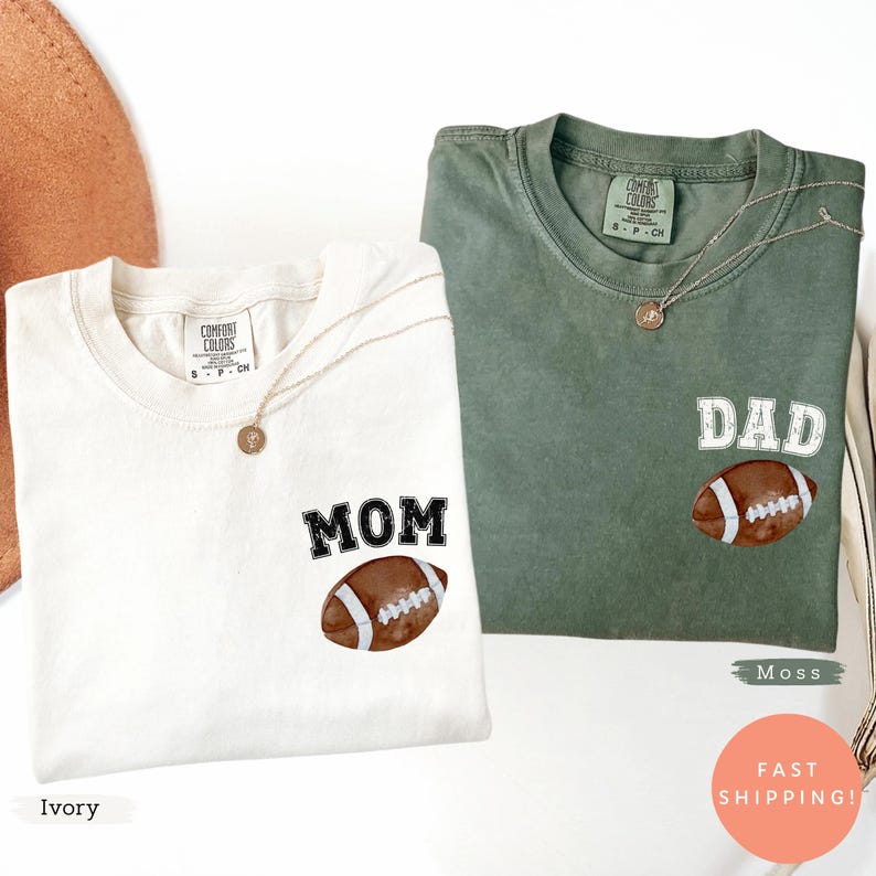 May include: Two Comfort Colors t-shirts, one ivory and one moss green, each with a football graphic and text. The ivory shirt says "MOM" and the green shirt says "DAD". Both shirts have a gold necklace.