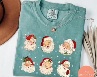 May include: Light green t-shirt featuring multiple Santa Claus faces with red hats and white beards. The shirt has a Comfort Colors tag and a silver necklace. The words "Light Green" are at the bottom left.