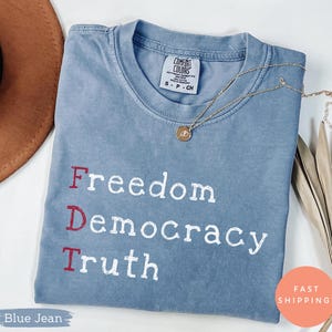 Könnte beinhalten: Ein blaues Comfort Colors T-Shirt mit den Wörtern "Freedom Democracy Truth" in Weiß und Rot aufgedruckt. Eine goldene Halskette mit Anhänger liegt auf dem Shirt. Das Shirt ist gefaltet und wird mit einem braunen Hut und getrockneten Blättern präsentiert.