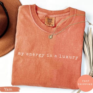 Puede incluir: Una camiseta de color coral con la frase "my energy is a luxury" impresa en blanco. Un collar de oro con un colgante descansa sobre la camiseta. La camiseta tiene una etiqueta Comfort Colors.