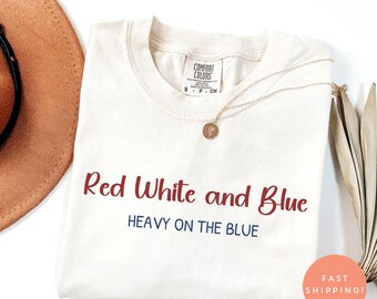 May include: Ivory-colored t-shirt with the text "Red White and Blue" in burgundy and navy blue. The shirt has a crew neck and short sleeves. A gold necklace with a pendant is draped across the shirt. A brown hat is partially visible.