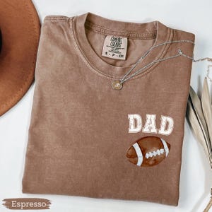 May include: Espresso-colored t-shirt with the word "DAD" in white block letters and a football graphic. The shirt has a crew neck and short sleeves. A silver necklace with a pendant is draped across the shirt.