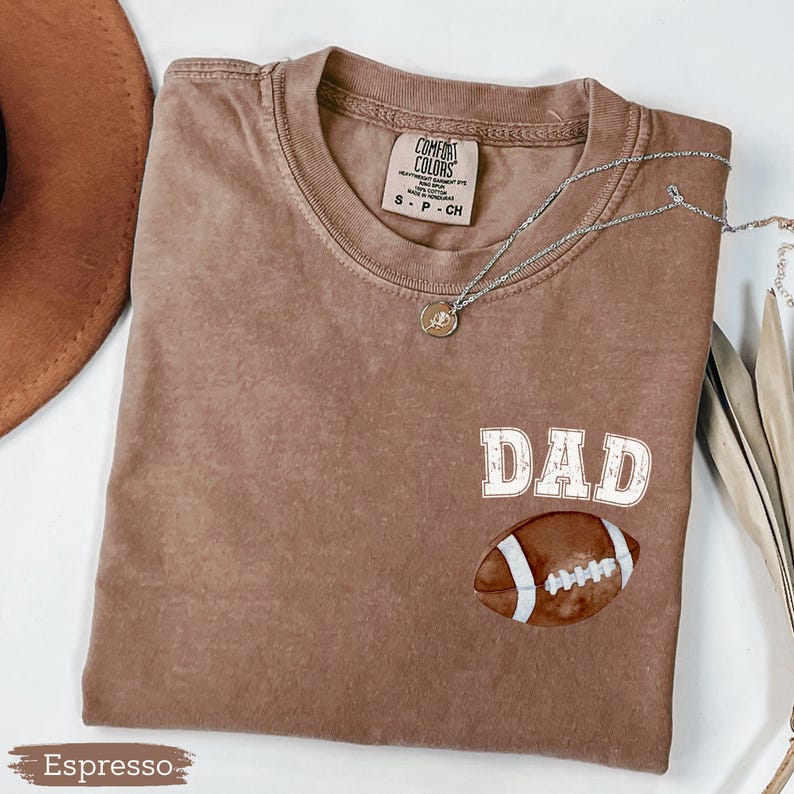 May include: Espresso-colored t-shirt with the word "DAD" in white block letters and a football graphic. The shirt has a crew neck and short sleeves. A silver necklace with a pendant is draped across the shirt.