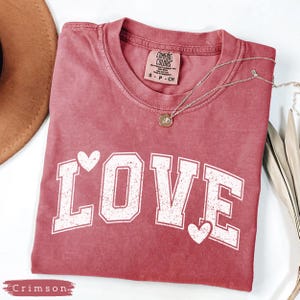 May include: Crimson-colored t-shirt with the word "LOVE" in white block letters and heart accents. A silver necklace with a pendant is draped across the shirt. The shirt is folded on a white surface.