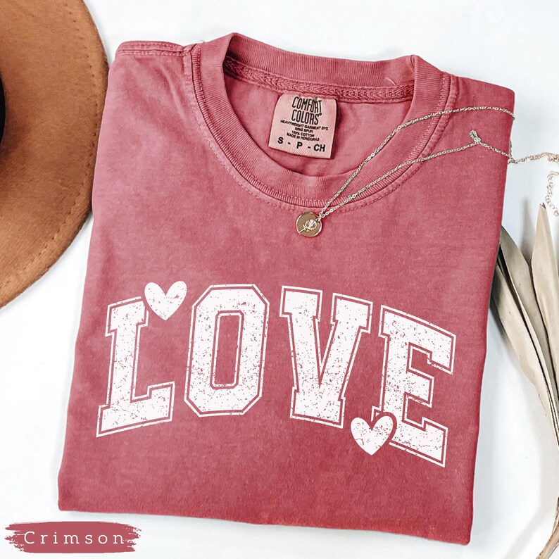 May include: Crimson-colored t-shirt with the word "LOVE" in white block letters and heart accents. A silver necklace with a pendant is draped across the shirt. The shirt is folded on a white surface.