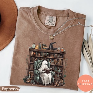 May include: Espresso-coloured t-shirt with a fantasy illustration of a ghost reading in a library. The design includes a bookshelf, a witch's hat, and potion bottles. The shirt has a crew neck and a small tag near the collar.