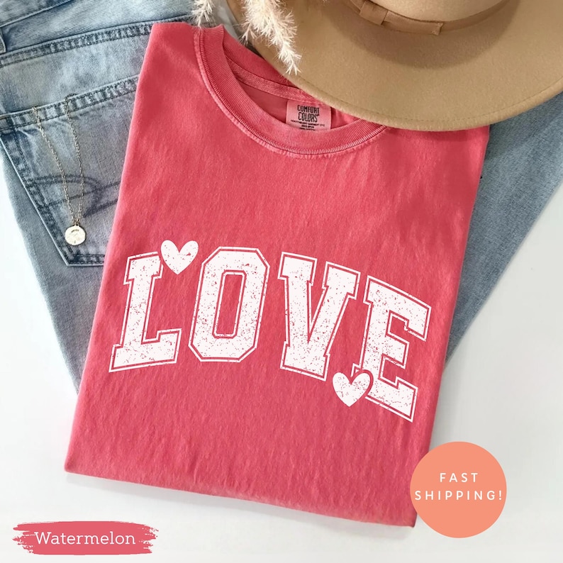 May include: A watermelon-colored t-shirt with the word "LOVE" in white, collegiate-style lettering. White hearts accent the letters. The shirt is folded on top of blue jeans, with a tan hat and necklace visible.
