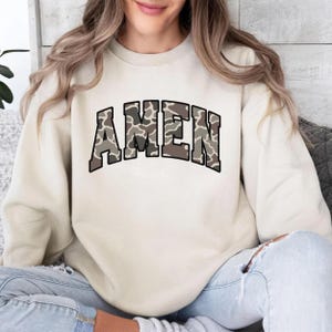 May include: A cream-colored sweatshirt with the word "AMEN" in large, block letters across the chest. The letters have a camouflage pattern in shades of brown and gray, outlined in black. The sweatshirt is worn by a person.
