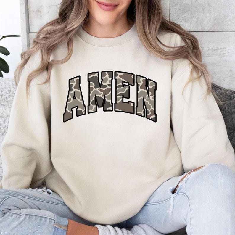 May include: A cream-colored sweatshirt with the word "AMEN" in large, block letters across the chest. The letters have a camouflage pattern in shades of brown and gray, outlined in black. The sweatshirt is worn by a person.