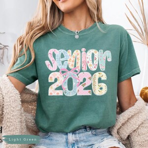 Może przedstawiać: Jasnozielona koszulka z napisem "Senior 2026" w patchworkowym wzorze. Tekst zawiera kwiatowe i kokardkowe akcenty. Koszulka jest w stylu casual z kr&oacute;tkim rękawem.