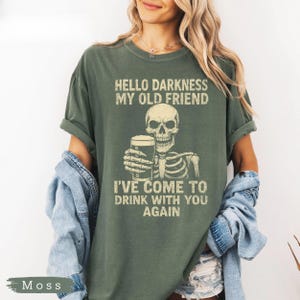 K&ouml;nnte beinhalten: Ein moosgr&uuml;nes T-Shirt mit einem Skelett, das ein Bierglas h&auml;lt. Der Text "HELLO DARKNESS MY OLD FRIEND I'VE COME TO DRINK WITH YOU AGAIN" ist in verblichener Schrift aufgedruckt. Das T-Shirt hat eine lockere Passform.