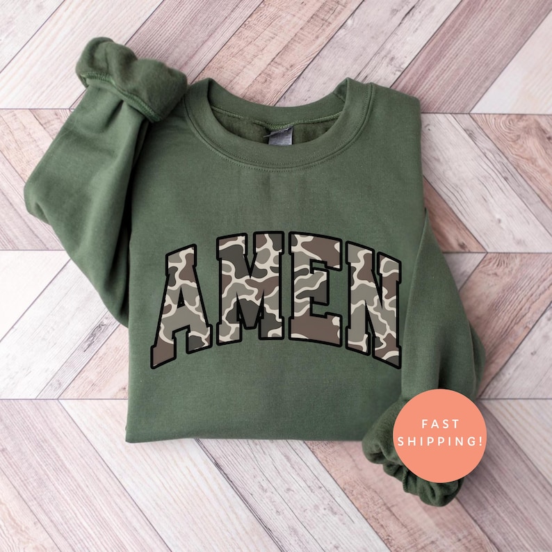 May include: An olive green sweatshirt with the word "AMEN" in a camouflage pattern across the chest. The sweatshirt has a crew neck and long sleeves. The text is outlined in a dark brown color.