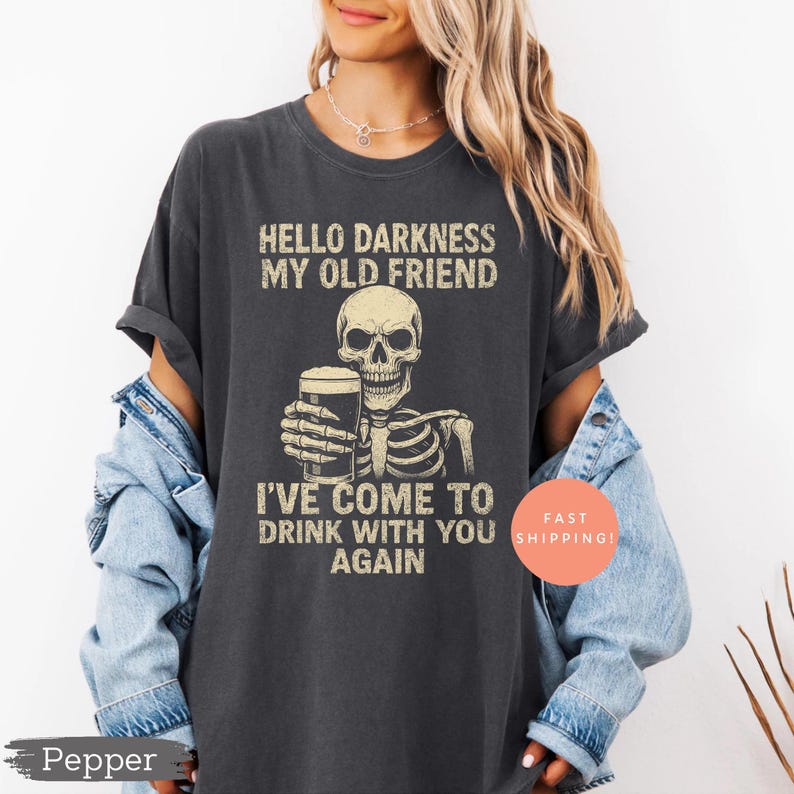K&ouml;nnte beinhalten: Dunkelgraues T-Shirt mit einem Skelett, das ein Bier h&auml;lt, und dem Text "HELLO DARKNESS MY OLD FRIEND I'VE COME TO DRINK WITH YOU AGAIN." Das Shirt hat eine lockere Passform.