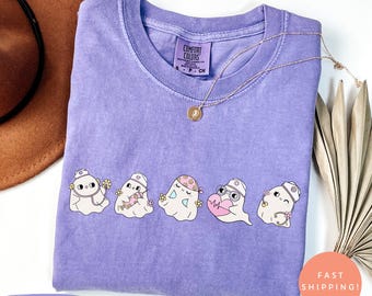 May include: A violet-coloured t-shirt with a crew neck and short sleeves. The shirt displays a graphic of five cartoon ghosts, each with a unique design, including flowers, a stethoscope, and a heart. A gold necklace is draped across the shirt.