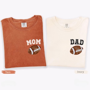 May include: Two folded t-shirts, one yam-colored and one ivory-colored. The yam shirt has the word "MOM" and a football graphic. The ivory shirt has the word "DAD" and a football graphic. Both shirts are made of a soft material.