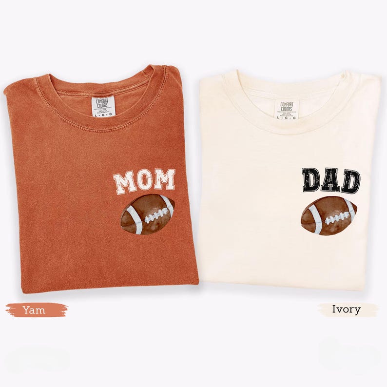May include: Two folded t-shirts, one yam-colored and one ivory-colored. The yam shirt has the word "MOM" and a football graphic. The ivory shirt has the word "DAD" and a football graphic. Both shirts are made of a soft material.