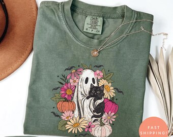 Könnte beinhalten: Moosgrünes T-Shirt mit einem Aufdruck, der ein weißes Gespenst zeigt, das eine schwarze Katze hält, umgeben von Blumen, Kürbissen und Fledermäusen. Das T-Shirt hat einen Rundhalsausschnitt und kurze Ärmel. Das Etikett zeigt "Comfort Colors".