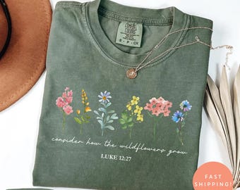 May include: Moss green t-shirt with a floral design and the text "consider how the wildflowers grow" and "LUKE 12:27". The shirt has a crew neck and short sleeves. A silver necklace with a pendant is on the shirt.
