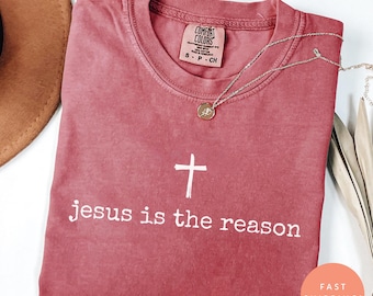 May include: Crimson-colored t-shirt with the text "jesus is the reason" and a white cross. A silver necklace with a round pendant rests on the shirt. The shirt has a Comfort Colors tag.