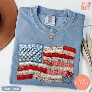 Peut inclure: Un t-shirt bleu jean Comfort Colors avec un drapeau américain en patchwork. Le drapeau comprend des étoiles blanches et des rayures rouges et à motifs floraux. Un collier en or avec un pendentif repose sur le t-shirt. Le t-shirt est étiqueté S-P-CH.