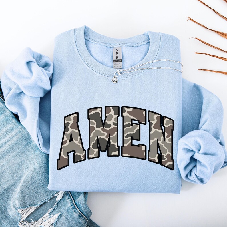 May include: A light blue sweatshirt with the word "AMEN" in a camouflage pattern across the chest. A silver necklace with a small pendant is draped across the sweatshirt. The sweatshirt is folded neatly on a white surface.