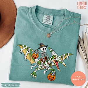 May include: A light green Comfort Colors t-shirt featuring a graphic of a skeleton wearing a witch hat riding a mummy-wrapped pterodactyl. The skeleton holds a jack-o'-lantern bucket, and the pterodactyl also carries a jack-o'-lantern bucket.