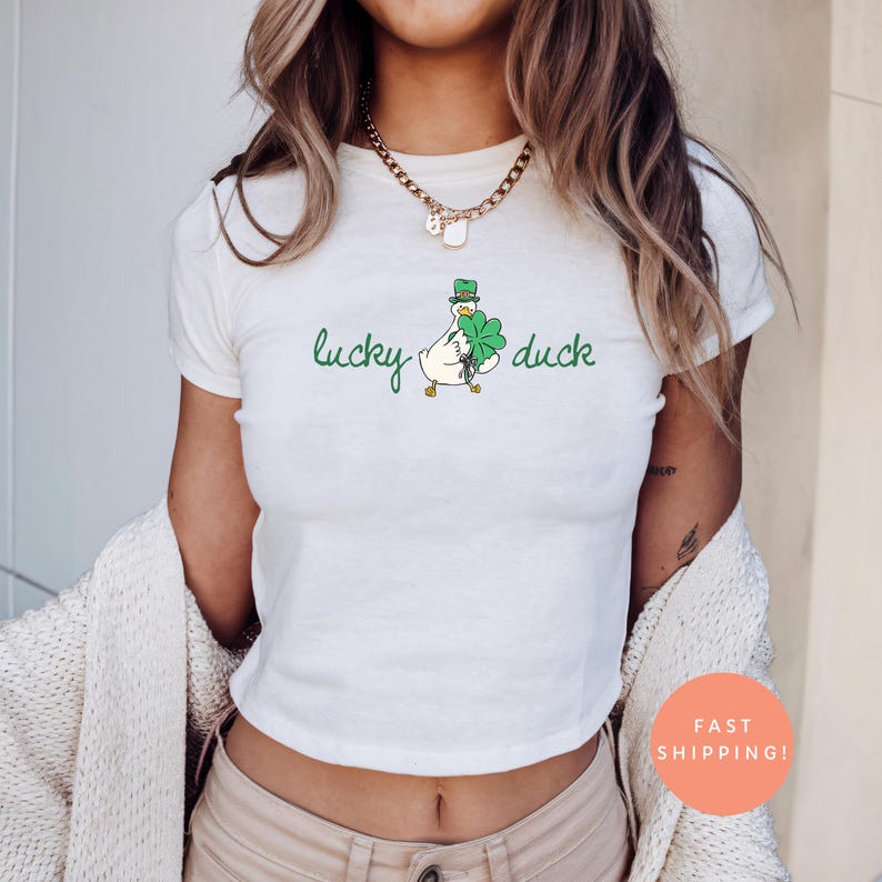 May include: Cream-coloured t-shirt with a green "lucky duck" design. The graphic shows a cartoon duck wearing a green hat and holding a four-leaf clover. The shirt is a cropped style.