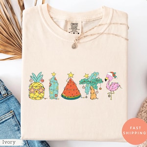May include: Ivory-colored t-shirt featuring a festive tropical design. The graphic includes a pineapple, surfboard, watermelon, palm tree, and flamingo, all decorated with Christmas lights and ornaments. The shirt has the text "Comfort Colors" on the tag.