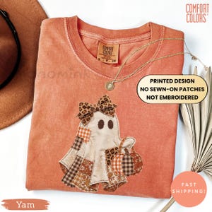 May include: A burnt orange t-shirt featuring a printed graphic of a ghost wearing a leopard print bow and holding a pumpkin. The ghost has a patchwork design with various brown and orange patterns. The shirt includes a Comfort Colors brand tag.