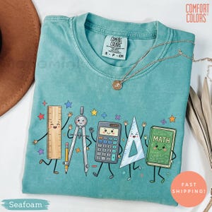 May include: A seafoam green crewneck t-shirt featuring a graphic print of five anthropomorphic school supplies including a ruler, compass, calculator, triangle, and a book labeled MATH, all with smiling faces and thin black limbs.