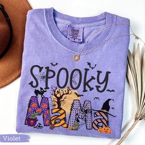 May include: A violet Comfort Colors t-shirt with the words "Spooky Mama" in black, orange, and purple, decorated with Halloween-themed graphics. A gold necklace and a brown hat are visible.
