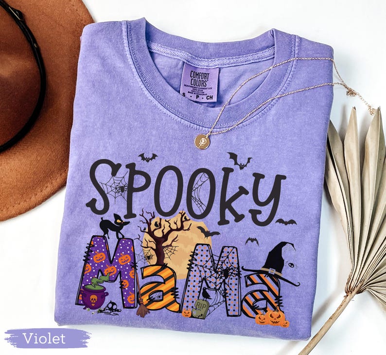 May include: A violet Comfort Colors t-shirt with the words "Spooky Mama" in black, orange, and purple, decorated with Halloween-themed graphics. A gold necklace and a brown hat are visible.