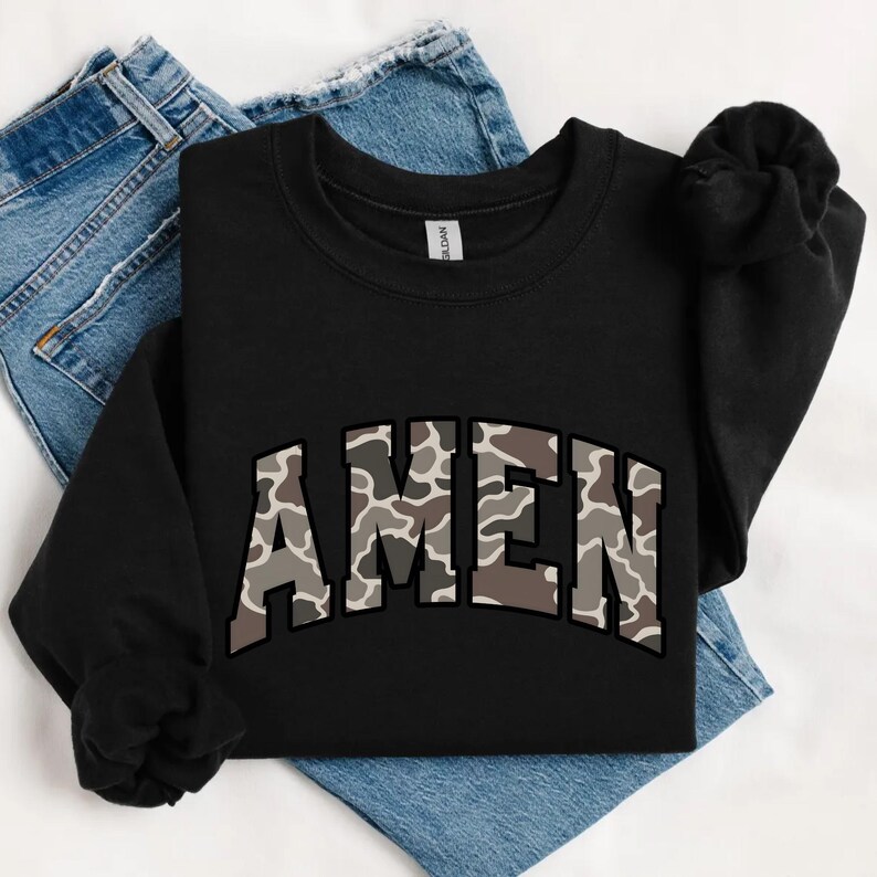 May include: Black crewneck sweatshirt with the word "AMEN" in a camouflage pattern across the chest. The sweatshirt is folded on top of a pair of blue jeans.